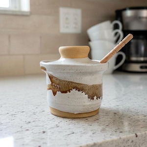 May include: A handmade ceramic butter dish with a lid and a wooden spoon. The dish is white with brown accents and a tan lid. The spoon is inserted into the dish. The butter dish is on a countertop.