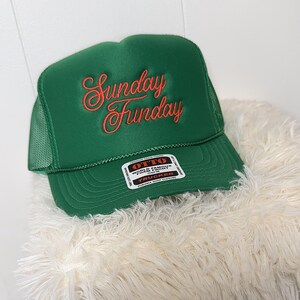 May include: A green trucker hat with the words "Sunday Funday" embroidered in a cursive, coral-colored font. The hat has a mesh back and a snap closure. A white tag with black text is visible on the front.