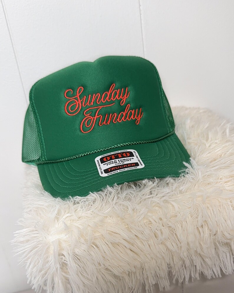 May include: A green trucker hat with the words "Sunday Funday" embroidered in a cursive, coral-colored font. The hat has a mesh back and a snap closure. A white tag with black text is visible on the front.