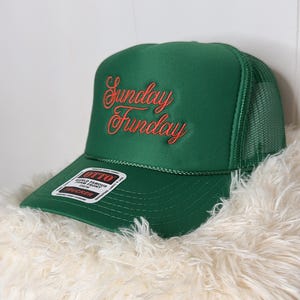 May include: A green trucker hat with the words "Sunday Funday" embroidered in red script. The hat has a mesh back and a curved brim. A white and black label is on the brim.