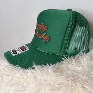 May include: Green trucker hat with the words "Sunday Funday" embroidered in red script. The hat features a mesh back and a curved brim. A tag is attached to the side of the hat.