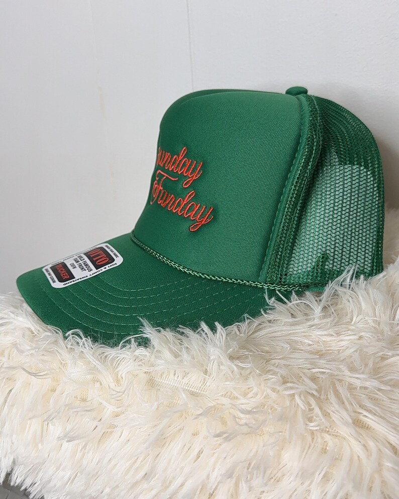 May include: Green trucker hat with the words "Sunday Funday" embroidered in red script. The hat features a mesh back and a curved brim. A tag is attached to the side of the hat.