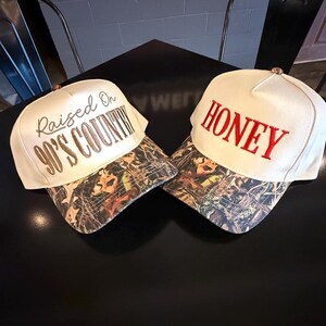 May include: Two white baseball caps with camouflage visors are displayed. One cap has "Raised On 90's Country" embroidered in brown, and the other says "HONEY" in red. The caps are on a black surface.