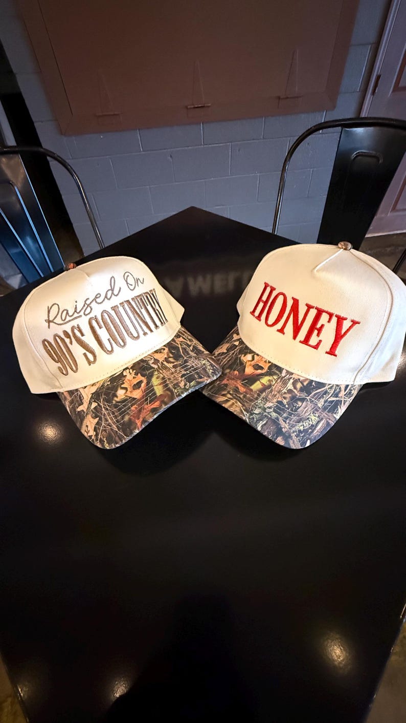 May include: Two white baseball caps with camouflage visors are displayed. One cap has "Raised On 90's Country" embroidered in brown, and the other says "HONEY" in red. The caps are on a black surface.
