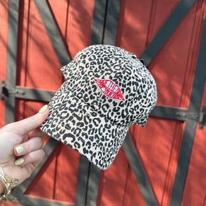 May include: A baseball cap with a leopard print pattern in black and tan. The cap features a red and white logo that reads "THE YEAR". The cap is held up in front of a red barn door.