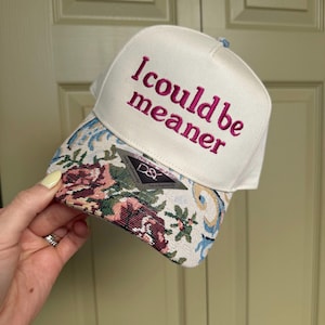 May include: White baseball cap with the embroidered phrase "I could be meaner" in pink. The brim features a floral tapestry design with pink, red, and green flowers. A small tag is visible on the brim.