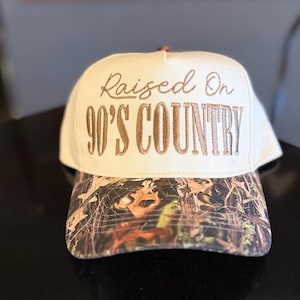 May include: Cream-colored baseball cap with the embroidered text "Raised On 90'S COUNTRY" in brown. The brim features a camouflage pattern in shades of brown, green, and orange. A classic accessory for country music fans.