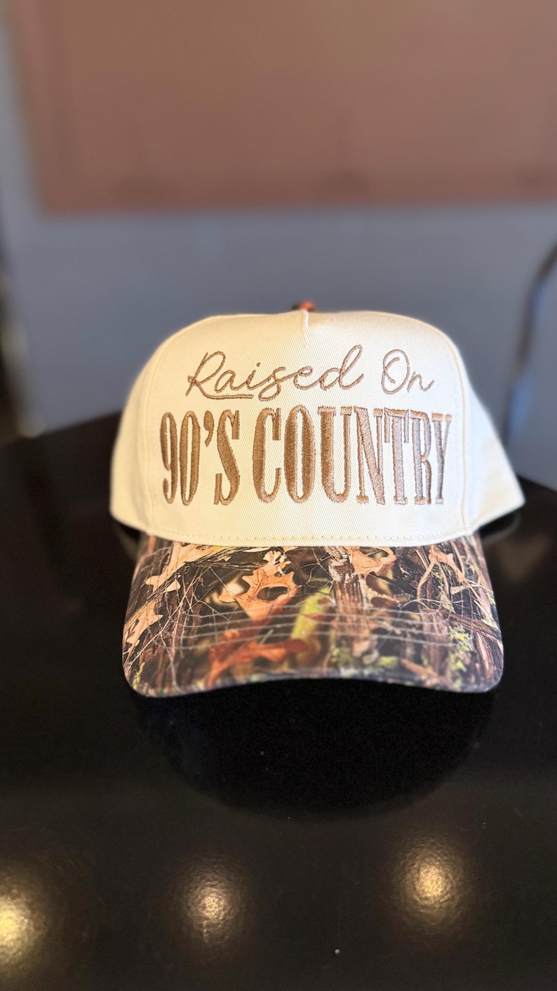 May include: Cream-colored baseball cap with the embroidered text "Raised On 90'S COUNTRY" in brown. The brim features a camouflage pattern in shades of brown, green, and orange. A classic accessory for country music fans.