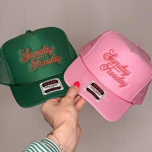 May include: A green and a pink trucker hat with the embroidered text "Sunday Funday" in a cursive font. The hats have a mesh back and a curved brim. Each hat has a white tag that reads "OTTO" and "Trucker".