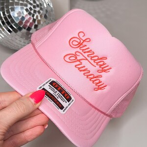 May include: A pink trucker hat with the embroidered text "Sunday Funday" in red script. The hat features a mesh back and a curved brim. A label on the side reads "World Famous Foam Front Trucker".