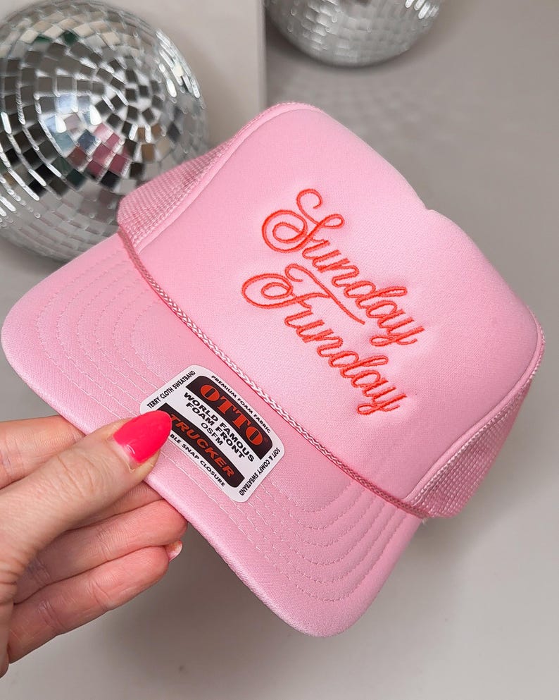 May include: A pink trucker hat with the embroidered text "Sunday Funday" in red script. The hat features a mesh back and a curved brim. A label on the side reads "World Famous Foam Front Trucker".