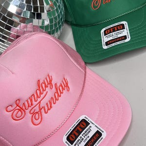 May include: Pink and green trucker hats with the text "Sunday Funday" embroidered in a cursive font. The pink hat has a matching pink mesh back, while the green hat has a green mesh back. Both hats have a "World Famous Foam Front" label.
