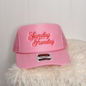 May include: Pink trucker hat with the text "Sunday Funday" embroidered in red script. The hat features a mesh back and a curved brim. A tag on the brim reads "OTTO World Famous Foam Front".
