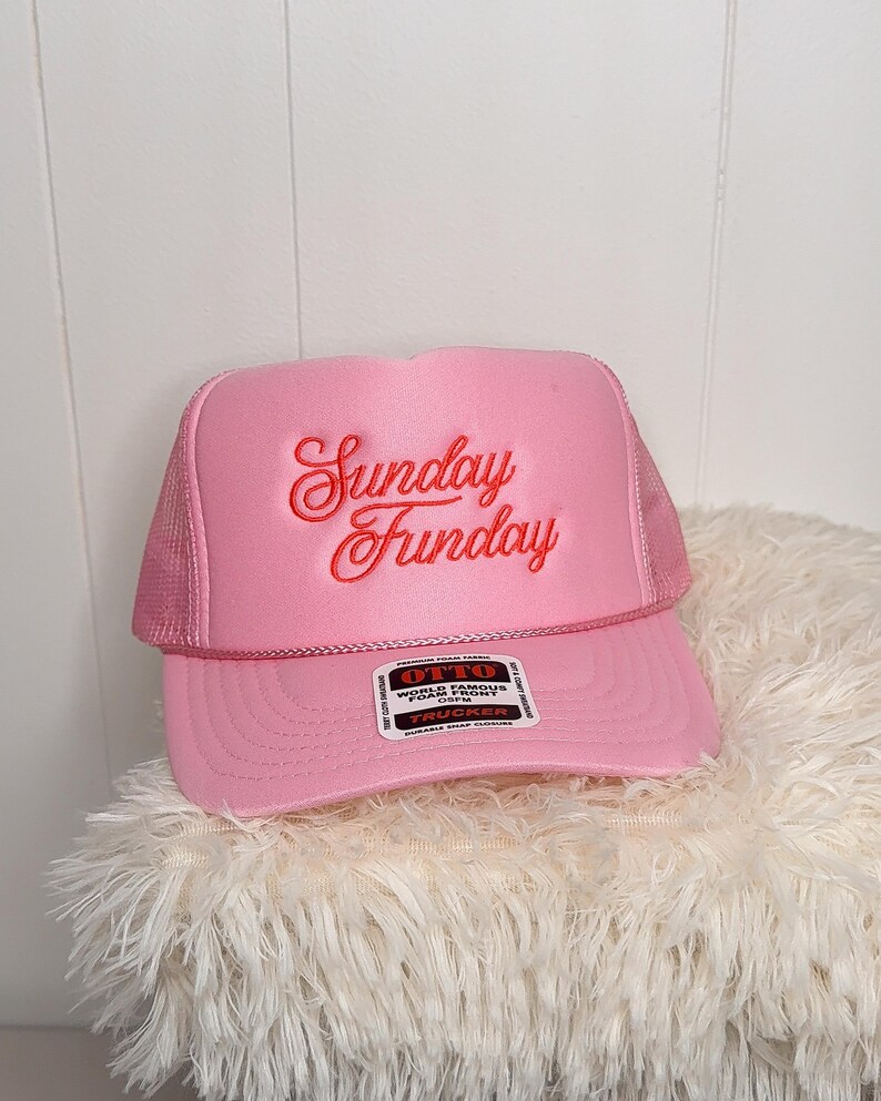 May include: Pink trucker hat with the text "Sunday Funday" embroidered in red script. The hat features a mesh back and a curved brim. A tag on the brim reads "OTTO World Famous Foam Front".