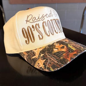 May include: A beige baseball cap with a camouflage brim. The cap features the embroidered text "Raised on 90's Country" in brown thread. The brim has a brown, orange, and green camouflage pattern.