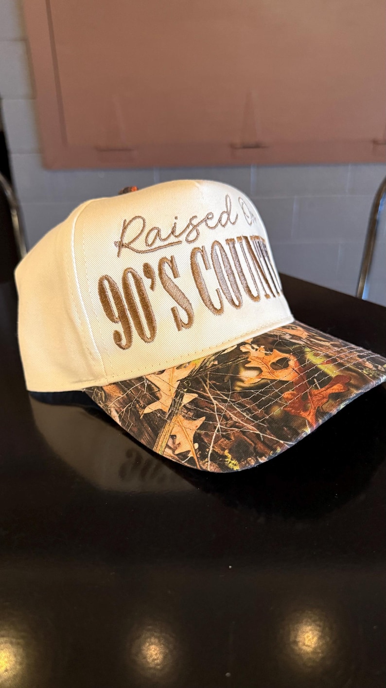 May include: A beige baseball cap with a camouflage brim. The cap features the embroidered text "Raised on 90's Country" in brown thread. The brim has a brown, orange, and green camouflage pattern.