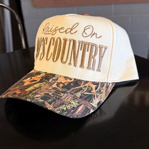 May include: Cream-colored baseball cap with the embroidered text "Raised On Country." The brim features a camouflage pattern with brown, green, and orange hues. The hat is sitting on a black surface.