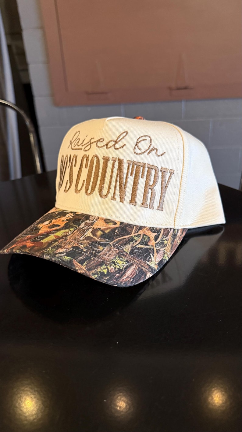 May include: Cream-colored baseball cap with the embroidered text "Raised On Country." The brim features a camouflage pattern with brown, green, and orange hues. The hat is sitting on a black surface.