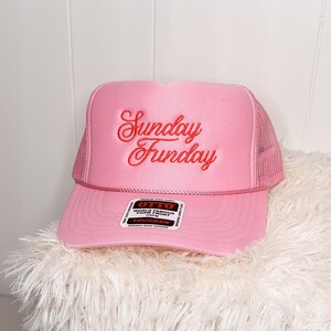 May include: Pink trucker hat with the words "Sunday Funday" embroidered in red script. The hat has a mesh back and a curved brim. The hat is sitting on a fluffy white surface.