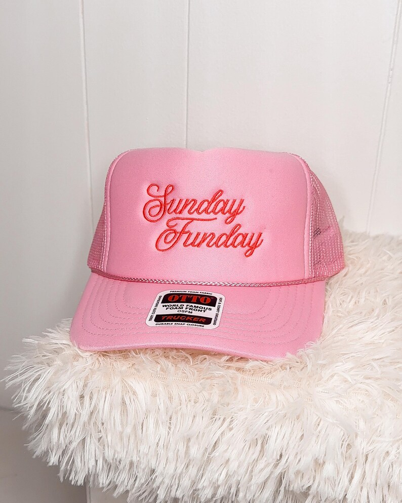 May include: Pink trucker hat with the words "Sunday Funday" embroidered in red script. The hat has a mesh back and a curved brim. The hat is sitting on a fluffy white surface.