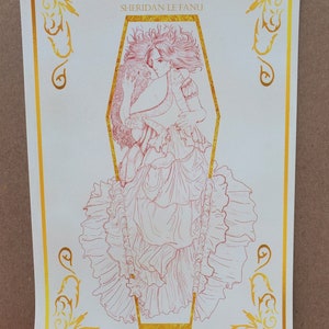May include: A white print with a gold border featuring a red line drawing of a woman in a coffin. The title "Carmilla" and the author "Sheridan Le Fanu" are printed in gold at the top of the print.