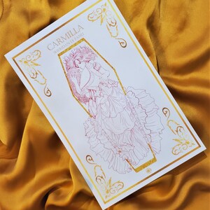 May include: A white card with gold foil accents featuring a line drawing of a woman in a coffin. The text "Carmilla Sheridan Le Fanu" is printed on the card.