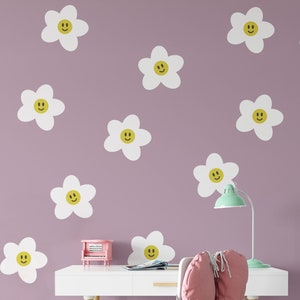 May include: A purple wall with white daisy decals with yellow smiley faces. The decals are arranged in a random pattern. There is a white desk with a pink chair and a small pink toy piano on the desk.