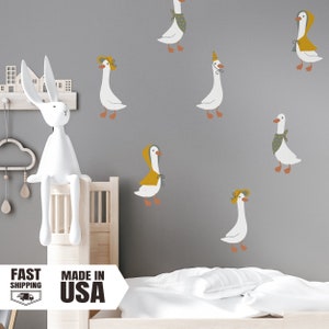 May include: A set of seven white goose wall decals with various accessories, including hats, scarves, and raincoats. The decals are on a gray wall, and there is a white rabbit figurine sitting on a wooden shelf.