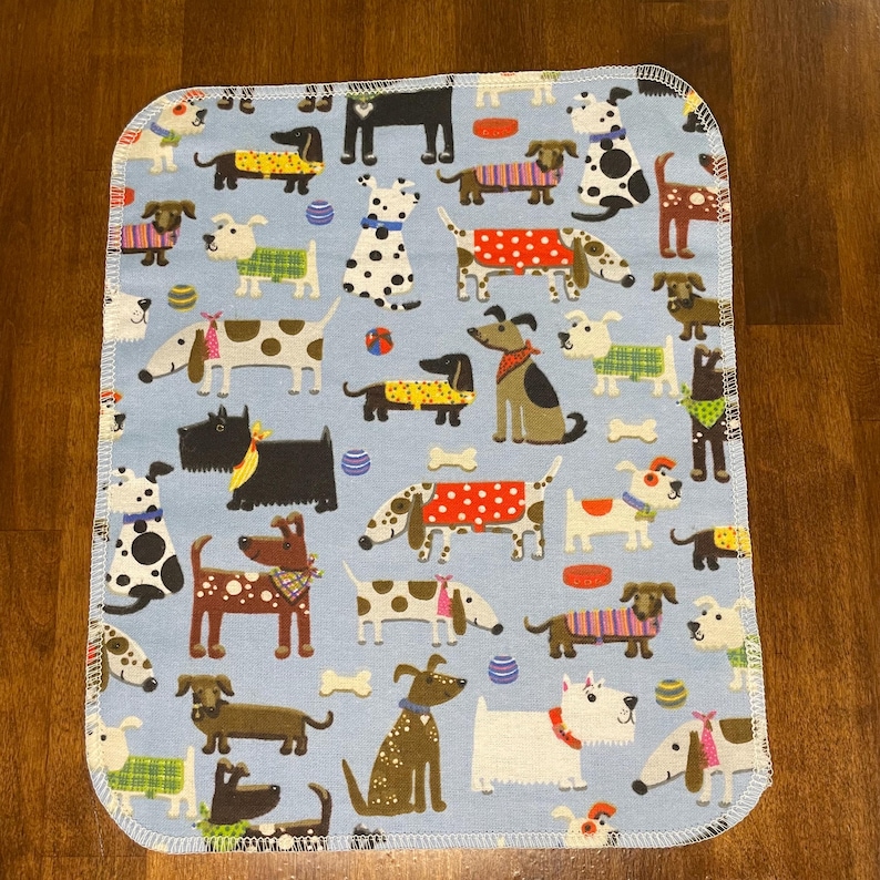 K&ouml;nnte beinhalten: Eine hellblaue, rechteckige Babydecke mit einem sich wiederholenden Muster verschiedener Hunderassen. Die Hunde sind in verschiedenen Farben und Outfits dargestellt, darunter Pullover und Bandanas. Die Decke hat einen wei&szlig;en, gen&auml;hten Rand.