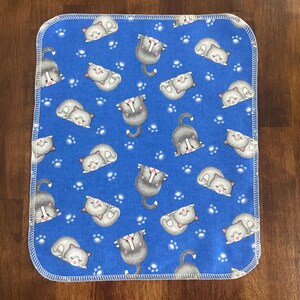 K&ouml;nnte beinhalten: Eine quadratische, blaue Babydecke mit einem sich wiederholenden Muster aus grauen Katzen und wei&szlig;en Pfotenabdr&uuml;cken. Die Katzen befinden sich in verschiedenen Posen, einige schlafen und andere stehen. Die Decke hat einen wei&szlig;en, gen&auml;hten Rand.