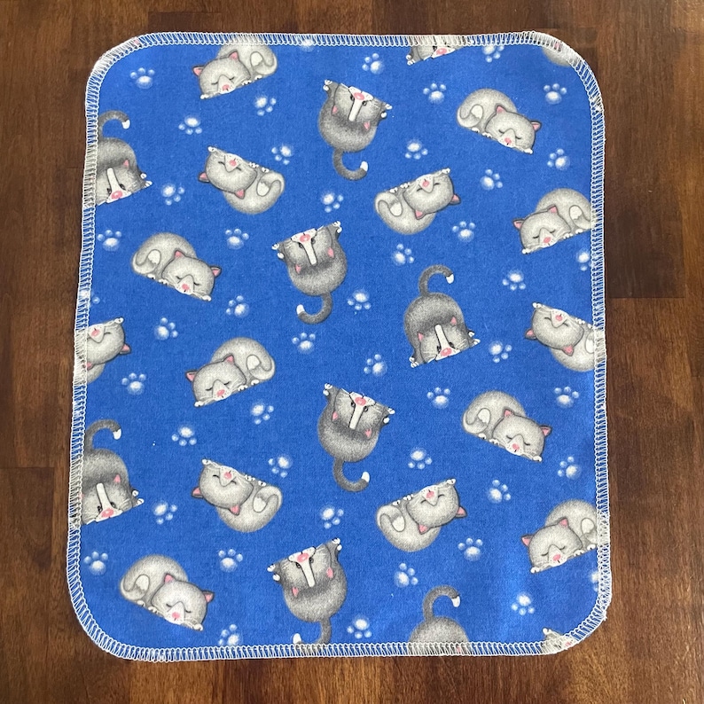 K&ouml;nnte beinhalten: Eine quadratische, blaue Babydecke mit einem sich wiederholenden Muster aus grauen Katzen und wei&szlig;en Pfotenabdr&uuml;cken. Die Katzen befinden sich in verschiedenen Posen, einige schlafen und andere stehen. Die Decke hat einen wei&szlig;en, gen&auml;hten Rand.