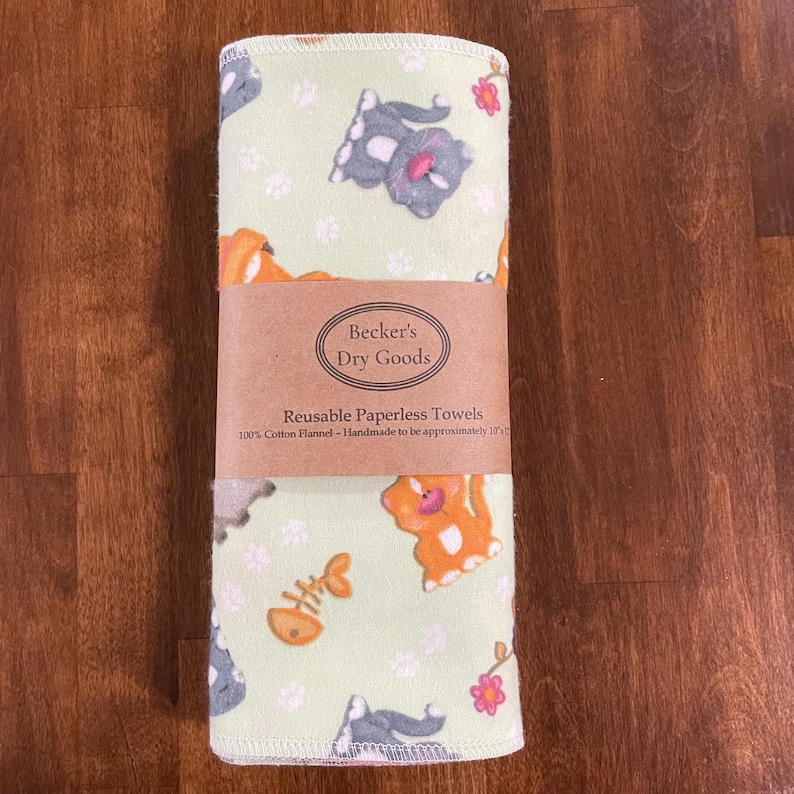 K&ouml;nnte beinhalten: Gerollte wiederverwendbare Papiert&uuml;cher mit hellgr&uuml;nem Hintergrund und einem Muster aus Cartoon-Katzen, Fischgr&auml;ten und Blumen. Die Handt&uuml;cher sind mit einem braunen Papierband mit der Aufschrift "Becker's Dry Goods" und "Reusable Paperless Towels" umwickelt.