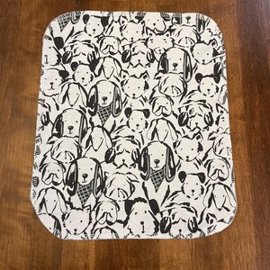 K&ouml;nnte beinhalten: Ein rechteckiges, cremefarbenes Baby-Spucktuch mit einem sich wiederholenden Muster aus schwarz-wei&szlig;en Hundegesichtern. Die Hunde haben verschiedene Ausdr&uuml;cke und Ohrformen. Die Kanten sind mit einer wei&szlig;en Naht versehen.