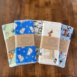 K&ouml;nnte beinhalten: F&uuml;nf wiederverwendbare Papiert&uuml;cher mit verschiedenen Mustern, darunter Katzen, Hunde und abstrakte Designs. Jedes Tuch ist mit einem braunen Papierband umwickelt, auf dem "Becker's Dry Goods" und "Reusable Paperless Towels" steht. Die T&uuml;cher sind ca. 25 x 30 cm gro&szlig;.