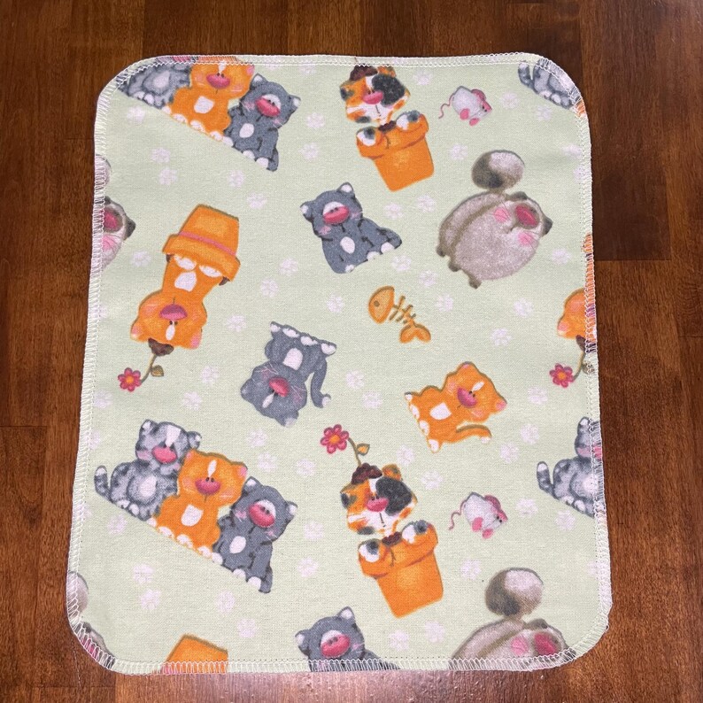 K&ouml;nnte beinhalten: Eine hellgr&uuml;ne Babydecke mit einem sich wiederholenden Muster aus Cartoon-Katzen in Orange, Grau und Schildpatt, zusammen mit kleinen rosa Blumen, Fischgr&auml;ten und M&auml;usen. Die Decke hat einen wei&szlig;en, gen&auml;hten Rand.