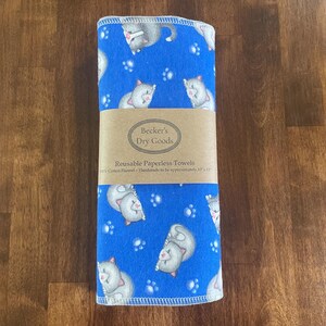 K&ouml;nnte beinhalten: Ein Stapel blauer, wiederverwendbarer Papiert&uuml;cher mit einem Muster aus grauen Katzen und wei&szlig;en Pfotenabdr&uuml;cken. Die T&uuml;cher sind mit einem braunen Papierband mit der Aufschrift "Becker's Dry Goods" und "Reusable Paperless Towels" umwickelt. Die T&uuml;cher sind ca. 25 cm x 30 cm gro&szlig;.
