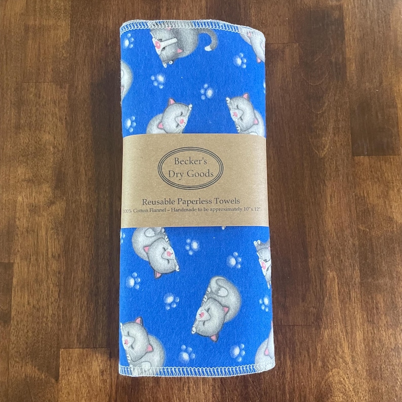 K&ouml;nnte beinhalten: Ein Stapel blauer, wiederverwendbarer Papiert&uuml;cher mit einem Muster aus grauen Katzen und wei&szlig;en Pfotenabdr&uuml;cken. Die T&uuml;cher sind mit einem braunen Papierband mit der Aufschrift "Becker's Dry Goods" und "Reusable Paperless Towels" umwickelt. Die T&uuml;cher sind ca. 25 cm x 30 cm gro&szlig;.