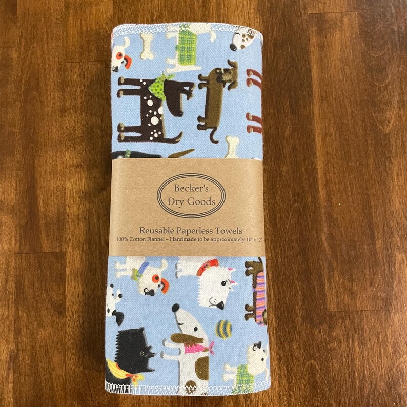 K&ouml;nnte beinhalten: Gerollte wiederverwendbare Papiert&uuml;cher mit hellblauem Hintergrund und einem verspielten Hundemuster. Die T&uuml;cher sind mit einem braunen Papierband mit der Aufschrift "Becker's Dry Goods" und "Reusable Paperless Towels" umwickelt. Die T&uuml;cher sind ca. 25 x 30 cm gro&szlig;.