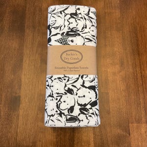 K&ouml;nnte beinhalten: Ein Stapel wiederverwendbarer Papiert&uuml;cher mit einem schwarz-wei&szlig;en Muster. Die T&uuml;cher sind mit einem braunen Papierband umwickelt, auf dem "Becker's Dry Goods" und "Reusable Paperless Towels" steht. Die T&uuml;cher bestehen aus 100 % Baumwollflanell.