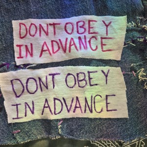 May include: Two white fabric patches sewn onto a blue denim background. The top patch says "Don't obey in advance" in pink lettering. The bottom patch says "Don't obey in advance" in purple lettering.