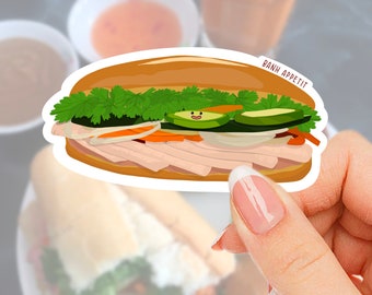 Może przedstawiać: Naklejka z kreskówkową ilustracją kanapki Banh Mi. Kanapka ma warstwy mięsa, warzyw i ziół, z napisem "BANH APPETIT" na górze. Naklejka jest trzymana przez dłoń z wypielęgnowanym paznokciem.