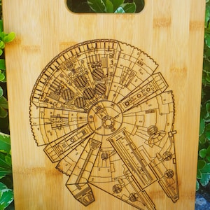 Puede incluir: Una tabla de cortar de madera con un diseño grabado con láser del Halcón Milenario de Star Wars. La tabla tiene un asa y está hecha de bambú.