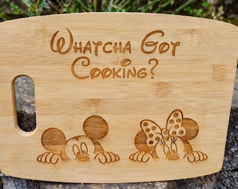 Puede incluir: Una tabla de cortar de madera con asa, con un diseño de Mickey Mouse y Minnie Mouse. La tabla tiene el texto "Whatcha Got Cooking?" grabado.