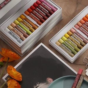 May include: Two open white boxes of Jieso Oil Pastels, each containing rows of colorful sticks. The pastels are in shades of red, orange, yellow, green, and blue. A drawing and orange flowers are also in the image.