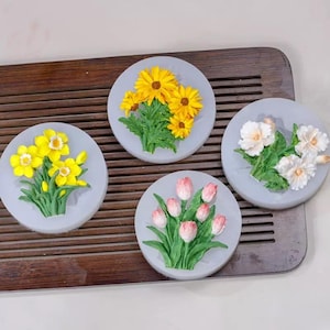 Könnte beinhalten: Vier graue, kreisförmige Untersetzer mit erhabenen Blumendesigns. Einer zeigt gelbe Narzissen, ein anderer gelbe Sonnenblumen, ein dritter weiße Blumen und der letzte rosa Tulpen. Die Blumen haben grüne Blätter und sind vor grauem Hintergrund.