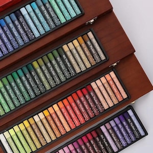May include: A wooden art box containing several trays of oil pastels. The pastels are organised by colour, including blues, greens, oranges, reds, and purples. Each pastel stick is labelled with the words "Oil Pastel".