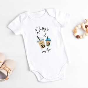 May include: White baby onesie with a graphic of two cartoon bubble tea cups and the text "Daddy's bes-tea".