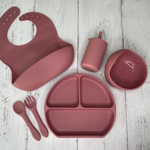 May include: A set of silicone baby feeding products in a dusty rose color. The set includes a bib, a bowl, a plate with three sections, a spoon, a fork, and a cup with a lid.