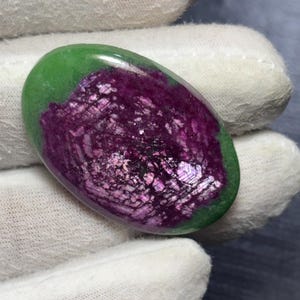 Puede incluir: Una gema ovalada con una vibrante combinación de colores. La mitad superior es de un púrpura profundo y brillante, mientras que la mitad inferior es de un verde intenso. La gema es sostenida por una mano enguantada.