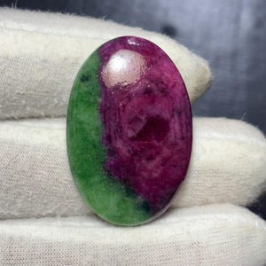 Peut inclure: Une pierre précieuse ovale polie, divisée en deux sections. Une section est vert vif, l'autre est magenta profond. La pierre est tenue par une main gantée de blanc.